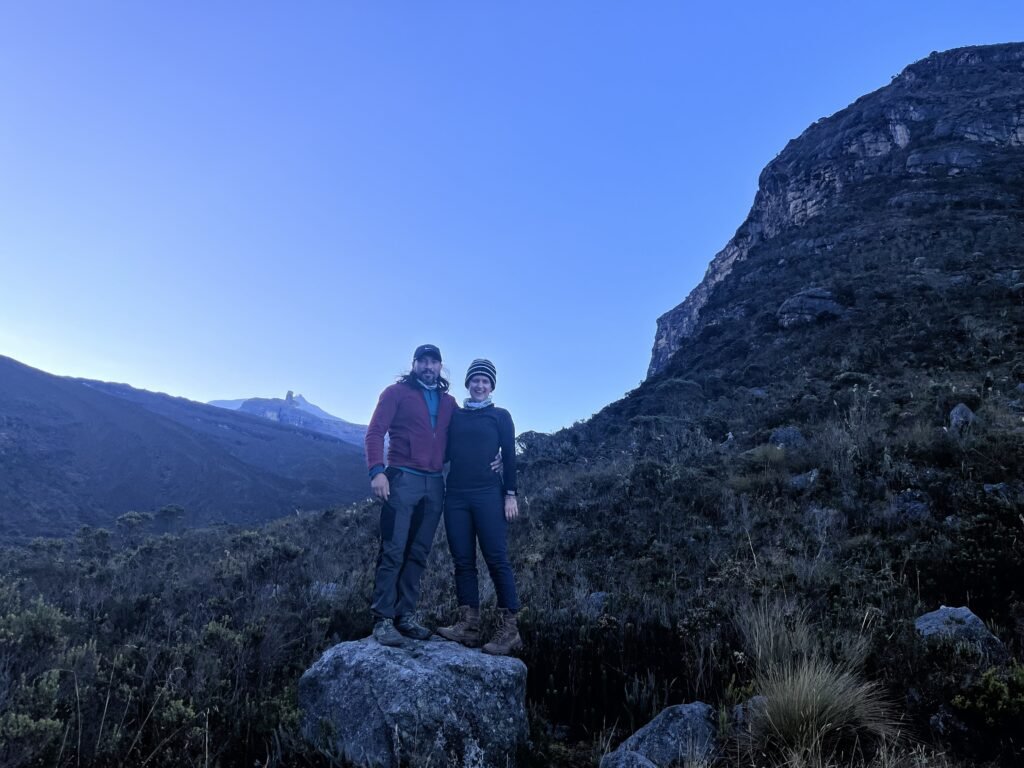 Couple standing ojn a rock in a colombian trail in cocyt national park with poles with the sun rising behind them and a snow peak behind them